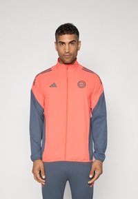 Heldere koraalkleurige jas met grijze mouwen, voorzien van een volle ritssluiting, hoge kraag en het Adidas-logo op de borst en mouw. Gevarieerde stof.