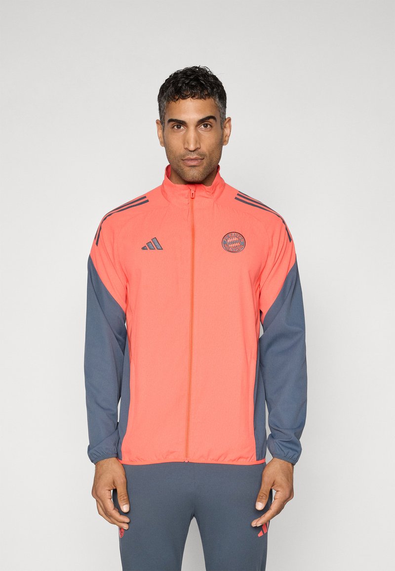 Heldere koraalkleurige jas met grijze mouwen, voorzien van een volle ritssluiting, hoge kraag en het Adidas-logo op de borst en mouw. Gevarieerde stof.