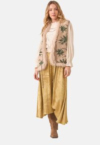 Chaleco de piel sintética marrón con bordado de hojas de palma, blusa cremosa texturizada, falda de satén dorado y botas marrones hasta la rodilla.