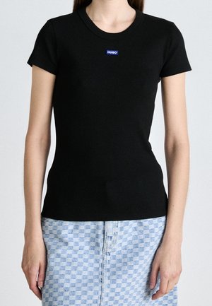 Kobieta ubrana w dopasowaną, czarną T-shirt z krótkim rękawem i małym niebieskim logo "HUGO" na piersi oraz jasnoniebieskie spodnie w kratę.