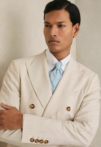 Blazer beige de doble botonadura con grandes botones de madera, usado sobre una camisa blanca y una corbata de lunares azul claro, con un corte entallado.