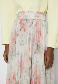 Jupe plissée fluide avec un doux motif floral dans des couleurs pastel, portée avec un haut à manches longues jaune pâle, montrant la taille et les mains.
