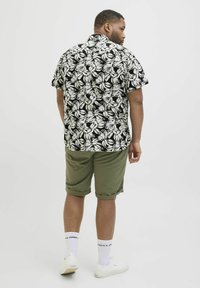 Schwarzes, kurzärmliges Hemd mit blühenden weißen Blättern, kombiniert mit olivgrünen, hochgerollten Shorts. Weiße Turnschuhe und Socken sind sichtbar.