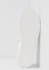 Semelle de basket blanche en caoutchouc texturé, avec un accent vert et noir. Le design se caractérise par des lignes épurées et une finition lisse.