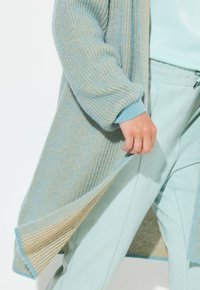 Offener Cardigan in Hellblau und Beige mit Rippenstruktur. Weicher Stoff, lange Ärmel und lockere Passform. Kombiniert mit hellblauen Hosen.