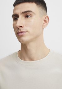 Neutralny beżowy t-shirt z okrągłym dekoltem, o gładkiej fakturze i minimalistycznym designie. Brak widocznych wzorów lub grafik.