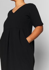 Schwarzes, kurzärmeliges Kleid mit V-Ausschnitt, geraffter Taille und Taschen. Hergestellt aus glattem Stoff mit lockerer Passform.