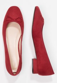Ballerines en daim rouge avec un bout arrondi, ornées d'un nœud à l'avant et d'un petit talon rectangulaire. Semelle intérieure en cuir lisse.