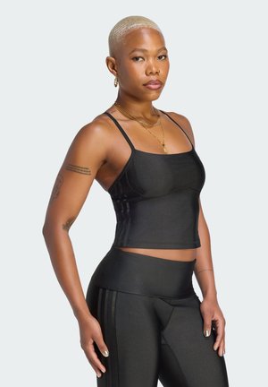 Donna con capelli corti biondi che indossa un crop top nero aderente e leggings, in piedi con le mani lungo i fianchi su sfondo uniforme.