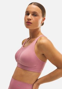 Sujetador deportivo rosa claro hecho de un material elástico con una textura suave. Presenta tirantes anchos, una banda de altura media y un sutil detalle de logo.