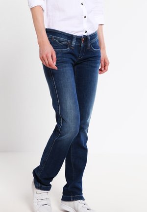 Personne portant un jean bleu foncé, une chemise blanche boutonnée et des baskets blanches, debout sur un fond clair uni.