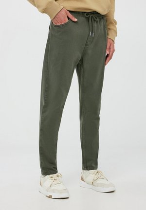 Persona con pantalones verde oliva con cordón y zapatillas blancas, con la mano en el bolsillo, de pie sobre un fondo claro liso.