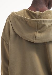 Sudadera con capucha de color verde oliva con un forro interior texturizado y un corte relajado. Costuras visibles a lo largo de la capucha y los hombros.