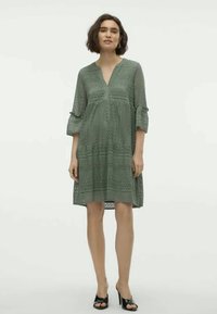 Žalia siuvinėta suknelė su V formos iškirpte ir trimis ketvirtadaliais varpo formos rankovėmis, puošta sudėtingais raštais ir laisva, plaukiančia siluetu.