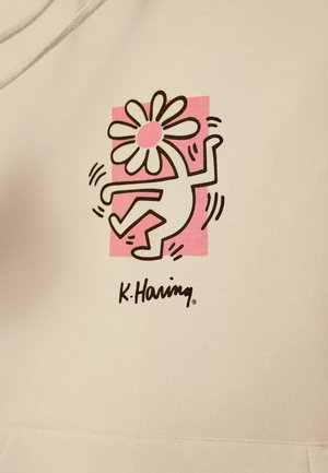Světle béžová látka má černý obrys tančící postavy s velkou květinovou hlavou, umístěnou na růžovém čtvercovém pozadí. Logo K. Haring je součástí.