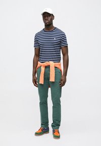 Μπλε ναυτικό και λευκό ριγέ βαμβακερό Τ-shirt, πορτοκαλί πλεκτό πουλόβερ δεμένο γύρω από τη μέση, πράσινο παντελόνι και πολύχρωμα αθλητικά παπούτσια με υφασμένα πέλματα.