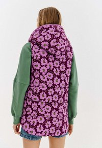 Kobieta w fioletowej kamizelce puchowej z różowym kwiatowym nadrukiem i kapturem, ubrana w zieloną bluzę i dżinsowe szorty, widziana z tyłu.