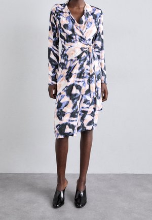 Femme portant une robe portefeuille longue aux genoux, à manches longues, avec un motif abstrait noir, blanc et pêche, et des chaussures à talons noires.
