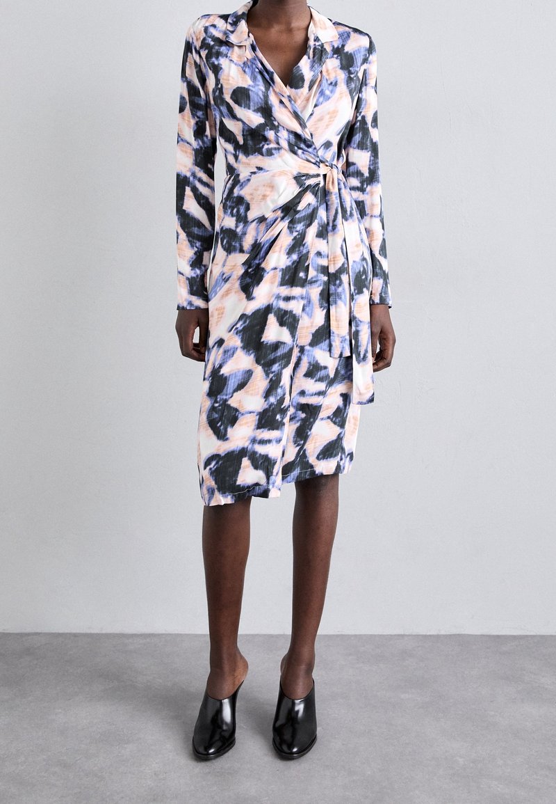 Femme portant une robe portefeuille longue aux genoux, à manches longues, avec un motif abstrait noir, blanc et pêche, et des chaussures à talons noires.