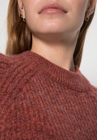 Gros plan sur le bas du visage et le cou d'une femme portant des boucles d'oreilles en or et un pull tricoté rustique texturé de couleur rouille.