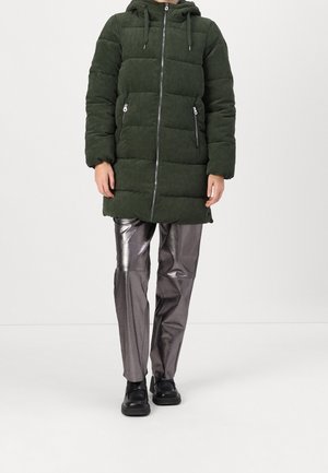 Personne portant un manteau matelassé à capuche vert foncé avec des poches zippées et un pantalon argenté métallique brillant avec des chaussures noires, debout sur un sol blanc.