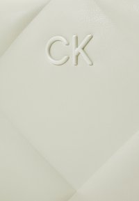 El material de cuero blanco presenta un patrón acolchado con un brillo sutil. El logo "CK" está grabado en un tono blanco similar.