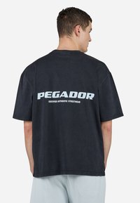 Mørk oversized t-skjorte laget av bomull, med fet hvit tekst som viser "PEGADOR" og mindre tekst nedenfor. Korte ermer og avslappet passform.
