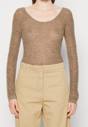 Femme portant un haut marron ajusté à manches longues en tissu duveteux, rentré dans un pantalon beige taille haute, debout devant un fond blanc uni.