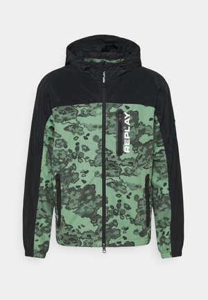 Leichte Zip-Jacke mit schwarzer Kapuze und Ärmeln, versehen mit einem grünen Camouflage-Muster auf dem Körper und einem auffälligen weißen Logo auf der Vorderseite.