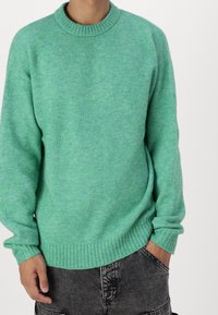 Zelený pletený svetr s žebrovaným kulatým výstřihem a dlouhými rukávy. Měkká textura, mírně volný střih, doplněný tmavě šedými šortkami.