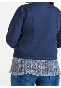 Persona con una chaqueta azul marino sobre una blusa blanca y azul con estampado, y jeans de mezclilla azules, vista desde atrás.