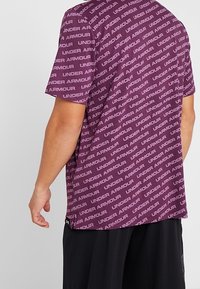 Lila t-shirt med ett mönster av texten "UNDER ARMOUR" över hela, rund hals, korta ärmar och en avslappnad passform.