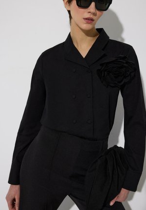 Ženska, oblečena v črno dvoredno srajco z veliko tkaninsko rožo na rami, usklajene črne hlače, drži črno torbico in nosi sončna očala.