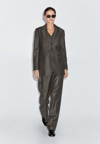 Vrouw die loopt in een oversized donker gestreept pak, vierkante zwarte zonnebril, zwarte schoenen tegen een effen lichte achtergrond.