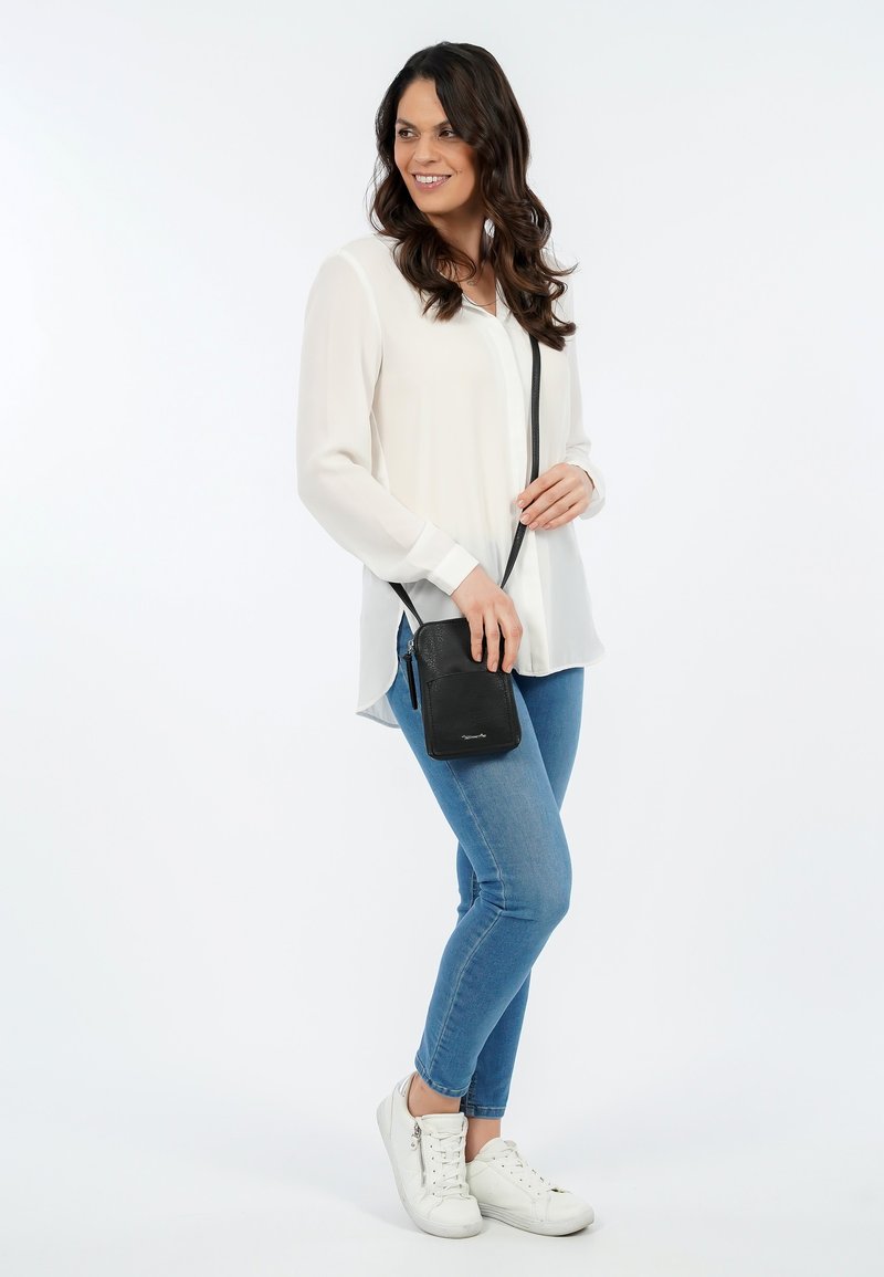 Borsa a tracolla nera in pelle, con tasca con zip e lunga tracolla, indossata con una camicia bianca e jeans blu. Sneakers bianche.