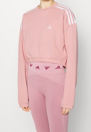 Persoon draagt een roze Adidas crop-sweatshirt met lange mouwen en witte strepen, gecombineerd met bijpassende hoge taille geribbelde leggings met een logo op de tailleband.