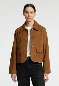 Brun ruskind cropped jakke med knaplukning foran, to forlommer og klassisk krave. Båret over en hvid skjorte og sorte bukser.