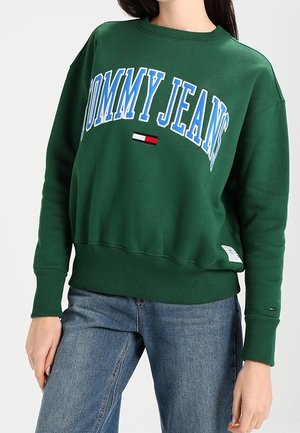 Osoba nosząca zieloną bluzę z napisem i logo "Tommy Jeans", w zestawieniu z niebieskimi dżinsami, stojąca na jednolitym tle.