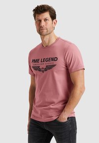 Camiseta de algodón rosa con un atrevido logo impreso en negro que dice "PME LEGEND AMERICAN CLASSIC" y un diseño de alas. Mangas cortas y cuello de barco.