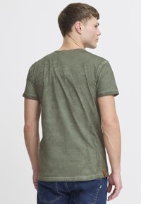 Oliven grøn kortærmet T-shirt med rund hals, tekstureret stof og opkrøllede ærmer. Har et lille orange mærke på venstre side.