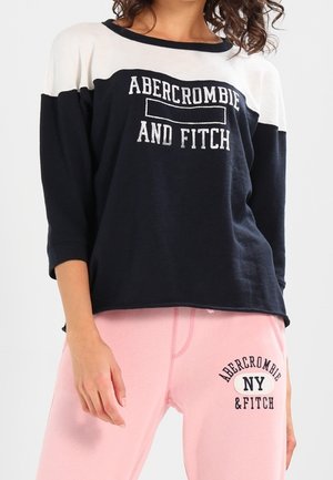 Kobieta ubrana w czarno-białą bluzę Abercrombie and Fitch oraz różowe spodnie z logo Abercrombie NY & Fitch na udzie.