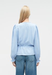 Lyseblå bluse med puffede ærmer, elastiske manchetter og en tætsiddende talje. Har en hakket halsudskæring og en glat tekstur. Kombineret med vidde bukser.