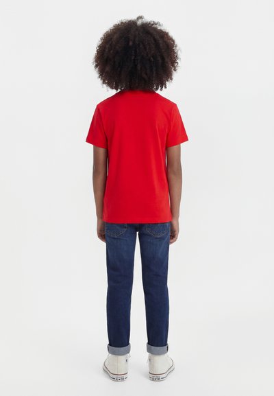 Child with curly hair wearing a red short-sleeve shirt, dark blue jeans, and white sneakers, standing facing away on a white background.
