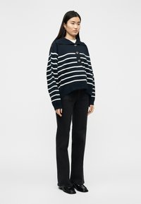 Femme debout portant un pull à boutons bleu marine rayé, un jean droit noir et des chaussures noires brillantes, sur un fond blanc.