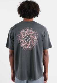 T-shirt en coton gris foncé avec un motif graphique spirale en rose et noir au dos, avec un patch logo sur l'épaule.