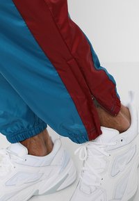 Person klädd i vita Nike-sneakers och blå träningsoverallbyxor med en vinröd rand och dragkedja vid ankeln, stående på en vit yta.