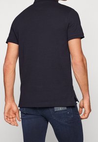 Marineblaues Poloshirt aus Baumwolle mit kurzen Ärmeln, einem umklappbaren Kragen und einem kleinen Logo am Saum, kombiniert mit dunklen Jeans.