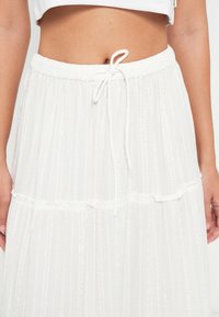 Jupe longue blanche à niveaux avec des rayures verticales texturées, taille élastique et fermeture à cordon. Tissu doux pour un confort léger.