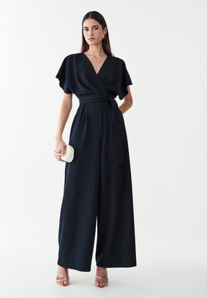 Vrouw met lang donker haar draagt een zwarte overslag jumpsuit en gouden oorbellen, houdt een kleine witte clutch vast en staat voor een witte achtergrond.