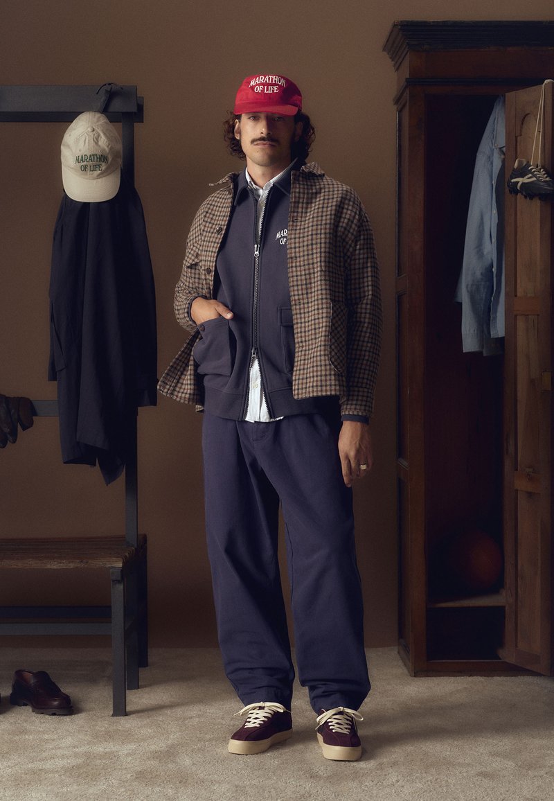 Model nosi smeđi karirani sako, tamnoplavu jaknu na zatvarač i odgovarajuće hlače. Crvena kapa s natpisom "MARATHON OF LIFE". Obuća kombinira boje maroon i krem.
